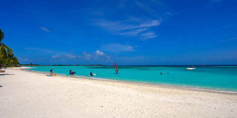 Fototapeta premium People enjoying water sports like windsurfing and paddleboarding on a beautiful white sand beach with vibrant turquoise water