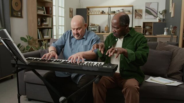 Front view shot of puzzled adult man getting music lesson at home and learning proper hand position for keyboard with male teacher helping Ideal for ads about adult education