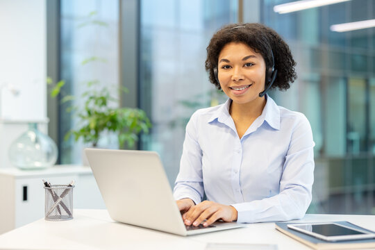 Portrait of a young African American woman in a suit sitting at an office desk with a laptop and looking at the camera with a smile