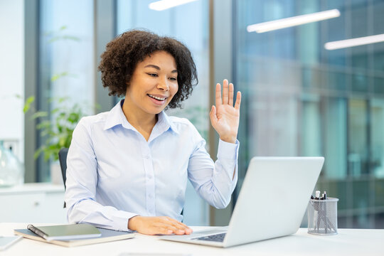African American young woman working in an office, sitting at a desk and talking on a laptop camera, waving