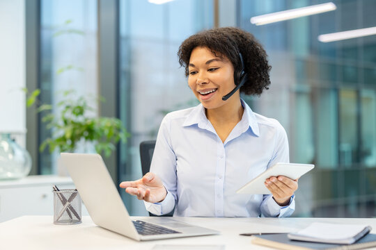 Smiling young African American woman sitting at a desk in the office wearing a headset, holding a tablet in her hand and talking on a video call