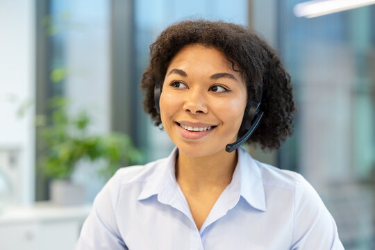 Close-up photo of a young African American woman in a headset sitting in an office at her workplace and looking to the side with a smile