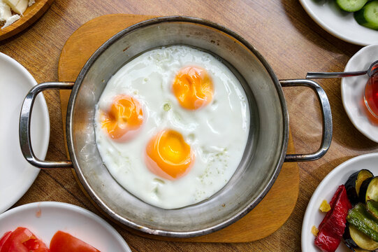 Cooking eggs in a metal pan with bright yolks alongside fresh vegetables and sauces on a wooden table