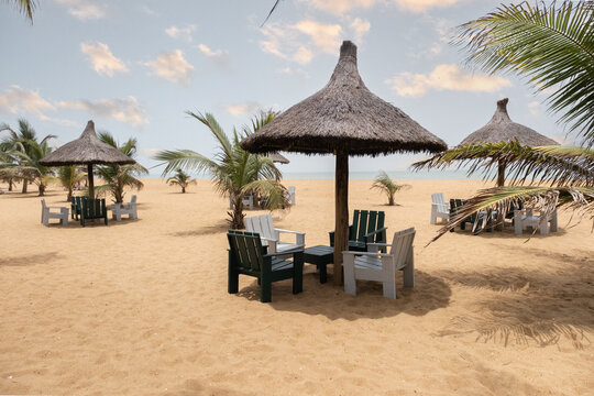 Ganvie, Benin - 14 April 2023: View of thatched umbrellas casting shadows on the golden beach, with palm trees swaying gently in the breeze, inviting relaxation.