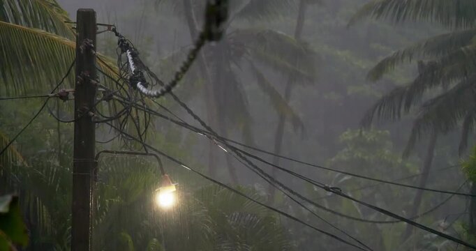 Glowing street lamp and tangled power lines under heavy tropical rain with palm trees in background Stormy wet season scene showing exposed electrical infrastructure and humid monsoon atmosphere