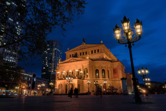 Frankfurt am Main, Germany - 13 April 2017: View of the historic Alte Oper, glowing golden against the deep indigo twilight, its grand facade contrasting with modern high-rises and ornate streetlamps.