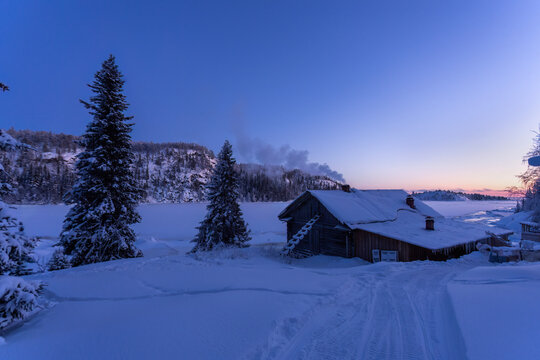 View of a rustic cabin dusted with snow stands peacefully under a fading winter sky, smoke rising gently, a scene of serene isolation, Kandalaksha, Murmansk Oblast, Russia.