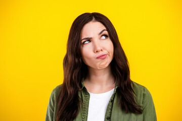Fototapeta premium Pensive young woman looking up with a puzzled expression against a bright yellow background, portrait conveying doubt, thinking and indecision in casual green shirt