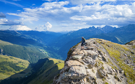 Aerial view of a lone hiker atop a rocky precipice, gazing into the endless expanse of verdant valleys and distant, jagged peaks, Nationalpark Hohe Tauern, Carinthia, Austria.