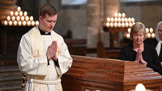 Priest and mourners praying with hands clasped at a wooden coffin during a solemn funeral service in a candlelit church