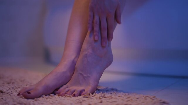 Close-up view of a woman's feet on a soft rug, gently massaging her tired and aching ankle, showing prominent varicose veins