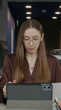 Vertical shot of young female office worker sitting at desk in modern workspace, reading business document and using digital tablet