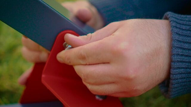 Firm grip adjusting metal frame connection without tool, emphasizing hand pressure and stability. Close detail of fingers securing joint on red bracket and dark beam. Assembly process concept