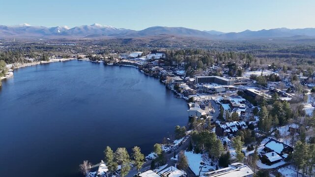 Aerial view of Mirror Lake's deep blue waters contrasting with snow-dusted buildings and distant white-capped mountains, Lake Placid, New York, United States.