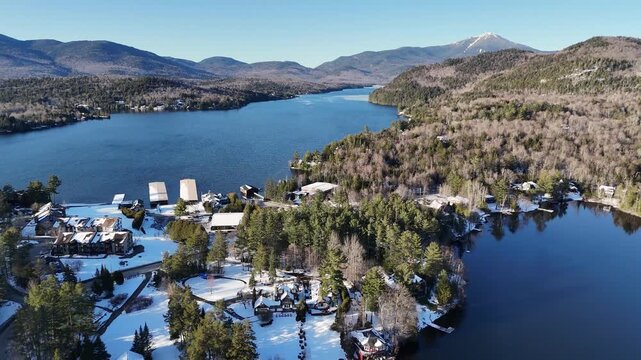 Aerial view of deep blue lake waters contrasting with crisp white snow on shorelines and rustic buildings, framed by dense forests and a distant majestic peak, Lake Placid, New York, United States.