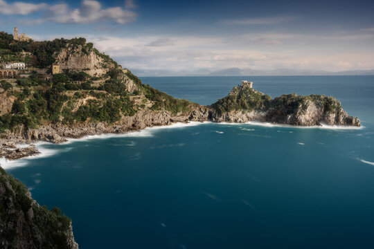 View of steep green cliffs meeting the deep blue sea, white waves churn below ancient towers, Conca dei Marini, Campania, Italy.