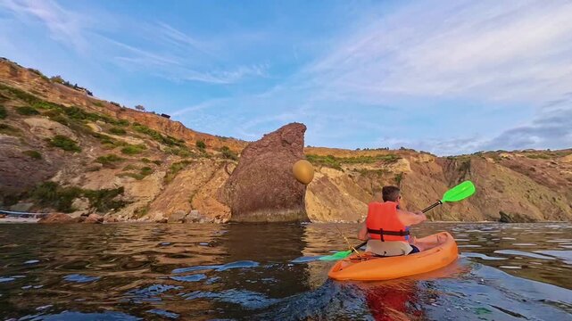 Kayak paddling sea adventure featuring a man rowing an orange boat along rocky cliffs with a gold balloon at sunset