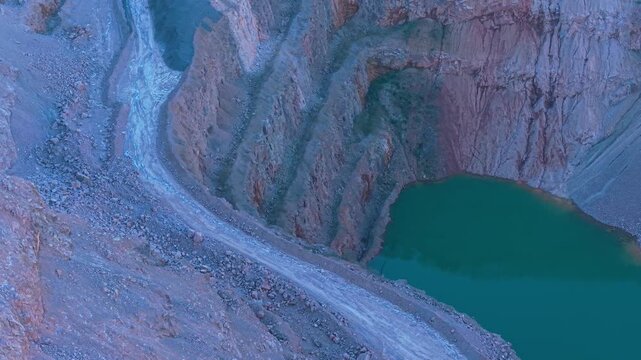 Serpentine quarry basin filling with green water between steep rock walls. Winding open pit excavation collecting water along layered mineral slopes. Developing artificial lake inside deep quarry