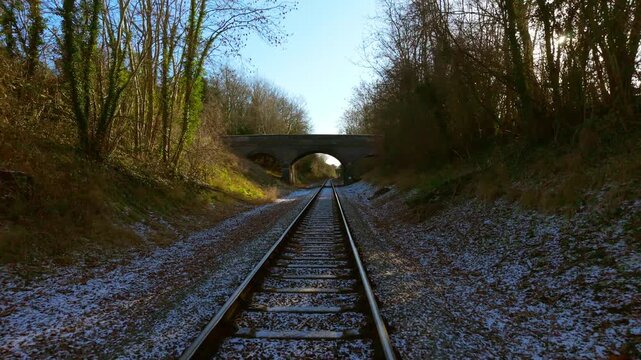 Passenger train moving forward along railway tracks toward stone bridge through winter cutting first person view. Commuter railcar advancing straight ahead on frosty rails between wooded embankments