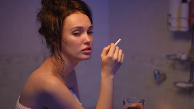Moody cinematic portrait of a beautiful young woman wrapped in a towel. She is relaxing in a bathroom, smoking a cigarette, and drinking wine