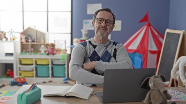 Man sitting at laptop smiling with arms crossed in school classroom with toys and chalkboard; teaching patience calm.