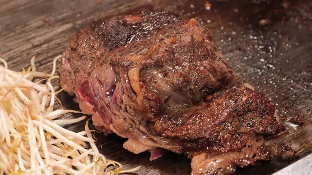 A professional chef skillfully slices a thick, marbled piece of premium Wagyu beef on a traditional Japanese teppanyaki iron griddle. The footage captures the sizzling meat, rich marbling, and expert 