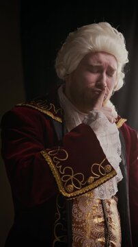 Vertical shot of young man in baroque embroidered coat and silver wig touching beard with discomfort, then raising finger to lips in silence gesture indoors