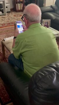 Anciano tercera edad con tel&eacute;fono m&oacute;vil sentado en sill&oacute;n del sal&oacute;n de casa haciendo scroll y deslizando pantalla smartphone leyendo noticias en celular