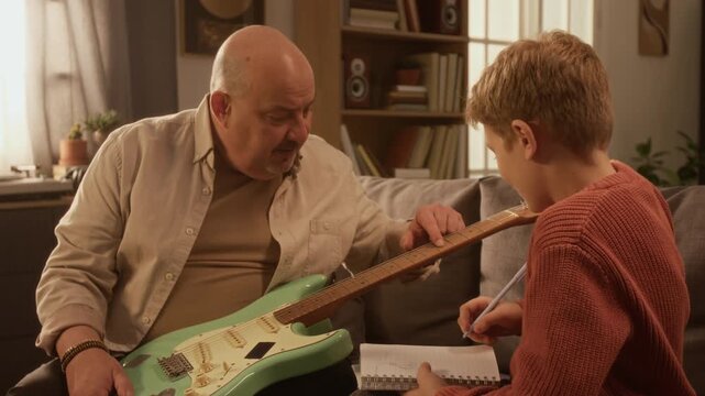 Medium shot of middle aged music teacher explaining electric guitar to young boy taking notes during music lesson at home scene lit by warm lighting Ideal for structured lesson promotion for kids