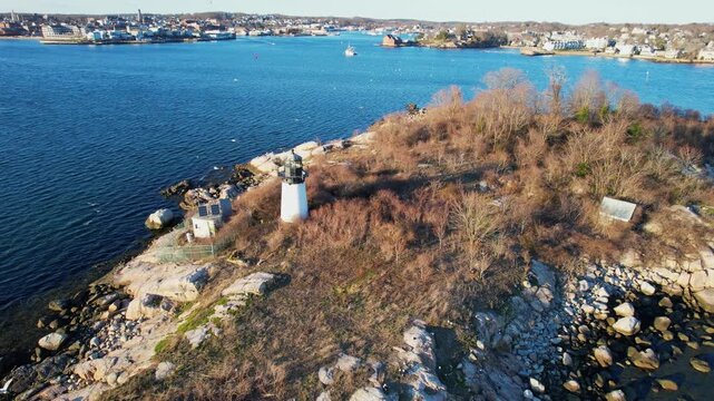 Aerial orbit of lighthouse in Gloucester, Massachusetts with rocky shoreline, blue harbor water and scenic coastal landscape creating classic New England seaside atmosphere on sunny day.