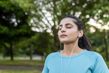 Fototapeta premium Young indian woman wearing earbuds and a blue t-shirt closing her eyes in concentration, meditating peacefully in a green park environment, finding relaxation and practicing mindfulness