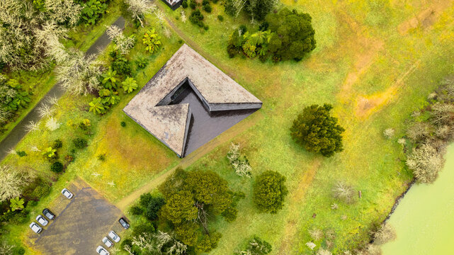 Aerial view of a striking geometric building with a central dark pool, nestled amidst vibrant green landscapes and a serene lake, Ponta Delgada, Azores, Portugal.