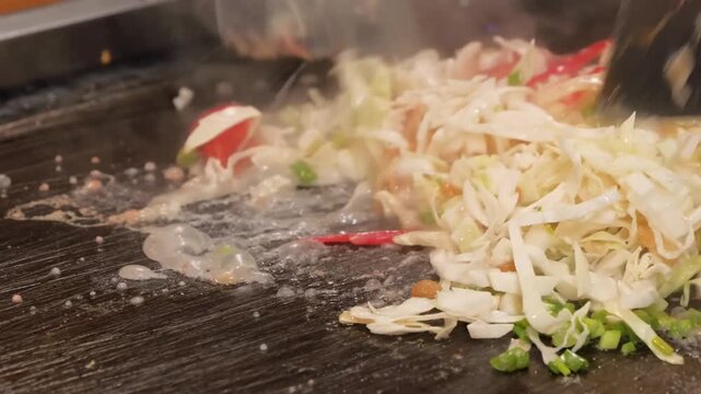 Close-up footage of a chef skillfully mixing and chopping fresh cabbage and ingredients to prepare Monjayaki, a savory pan-fried batter dish. This popular Tokyo street food is cooked on a hot iron tep