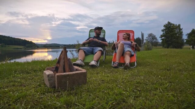 Couple sitting on outdoor chairs near campfire by calm lake at sunset, talking and enjoying peaceful camping atmosphere in natural setting of Savci Slovenia