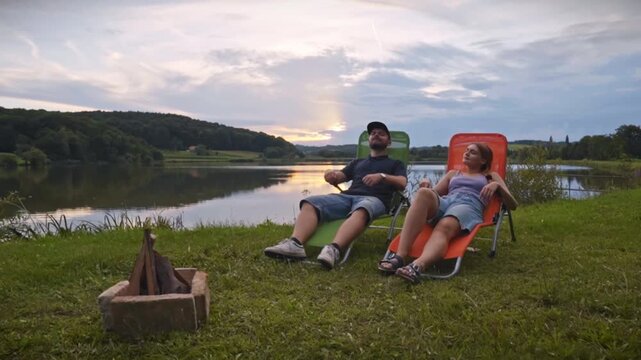 Couple sitting on outdoor chairs near calm lake with campfire at sunset, talking and enjoying peaceful camping atmosphere in natural setting of Savci Slovenia