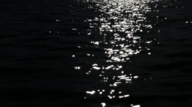 No harshness. A dark water surface at dusk with a faint, narrow, shiny trail of light reflecting off the slow ripples, sparkles barely visible against the dark blue water in the low ambient light.