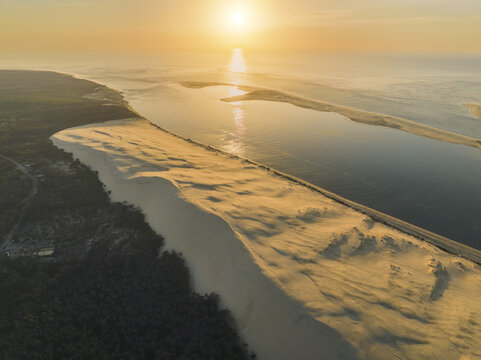 Aerial view of golden sands meeting the tranquil waters under a radiant sun, a coastal symphony of light and shadow at Dune du Pilat, La Teste de Buch, Nouvelle-Aquitaine, France.