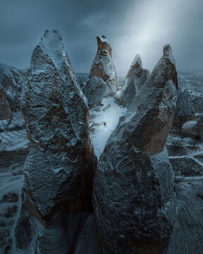 Aerial view of snow-dusted fairy chimneys, their rugged textures contrasting with soft white and a distant glow piercing the cloudy sky, Goreme, Turkey.