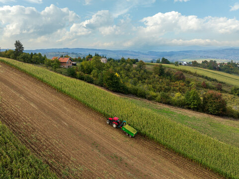 Aerial view of a vibrant red tractor harvesting golden green crops across textured brown fields under a wide, cloud-strewn sky, Tuzla, Bosnia and Herzegovina.