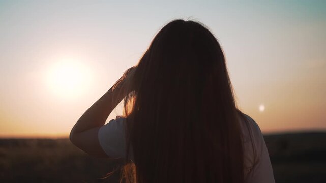 Woman touches hair during sunset with golden light outdoors. Female silhouette at sunset horizon. Woman with backlit hair at evening. Sunset creates silhouette while woman touches hair outdoors.