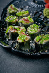 Sushi maki rolls topped with seaweed salad (chuka), sesame seeds, and balsamic glaze, served with ginger and wasabi on a dark plate © ismishko