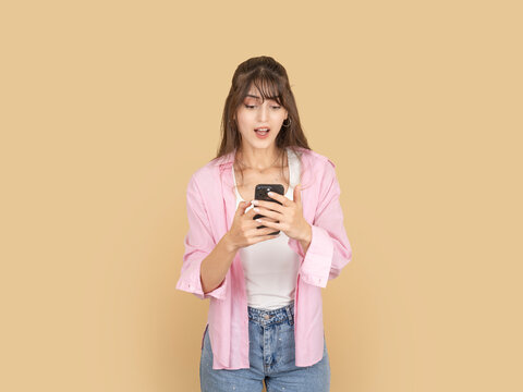 Surprised young woman using smartphone with open mouth expression, reacting to unexpected message. Isolated on beige background copy space, ideal for communication, news and digital reaction concepts.