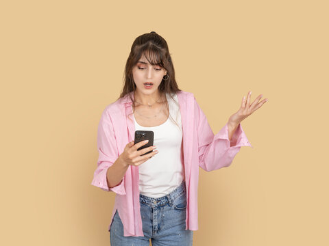 Confused young woman holding smartphone and gesturing with raised hand  reacting to message. Isolated on beige background with copy space, ideal for problem, communication or misunderstanding concept.