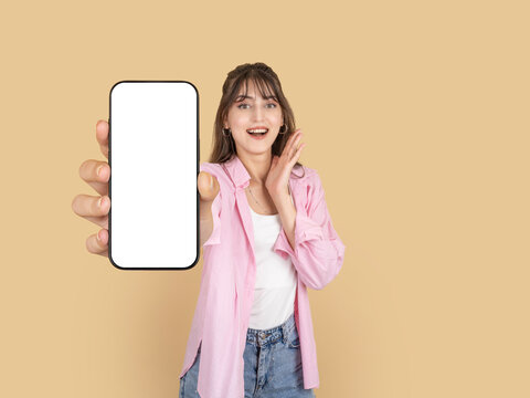 Excited young woman holding smartphone mock up, blank screen to camera, reacting surprise expression. Isolated beige background,  copy space, ideal for mobile app promotion and advertising concepts.