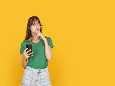 Thoughtful young woman holding smartphone and touching chin while looking aside copy space text area. Isolated on bright yellow background. Decision making or mobile usage concept idea image.