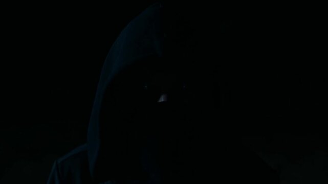 Slow zoom closeup of hooded figure with glowing eyes appearing and fading in dark urban alley at night