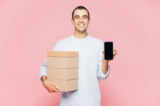 Young smiling middle eastern man he wears casual blue shirt hold in hands stack cardboard blank boxes delivery use mobile cell phone with blank screen workspace area isolated on plain pink background.