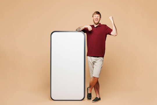Full size young smiling happy man wear red shirt stand at big huge blank screen mobile cell phone smartphone with workspace copy space mockup area isolated on light beige background Lifestyle concept
