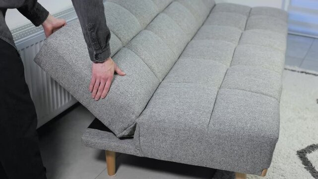 Man demonstrating a versatile grey sofa bed, transforming it into three different positions: fully flat for sleeping, semi-reclined for lounging, and upright for sitting in a modern interior.