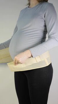 Vertical footage of pregnant woman adjusting a supportive maternity belt over her grey shirt to alleviate back pain and provide abdominal stability during pregnancy.